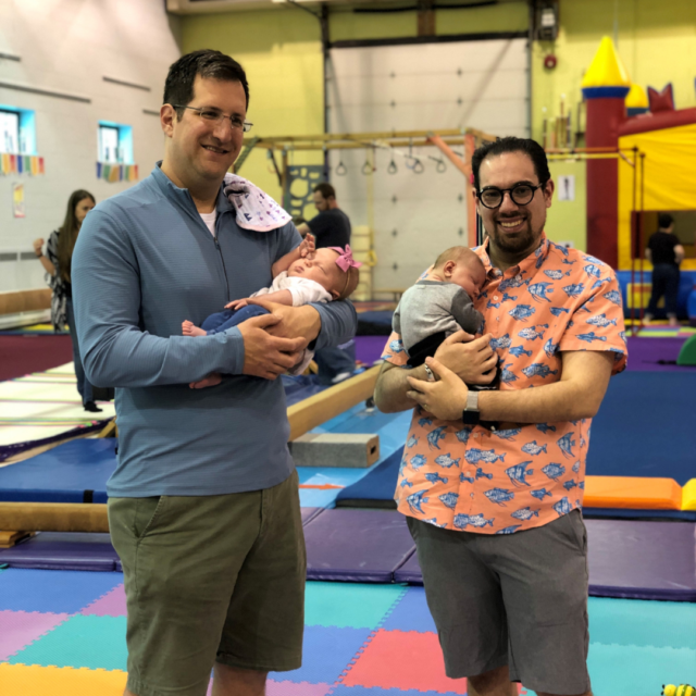 two dads holding babies in a gym