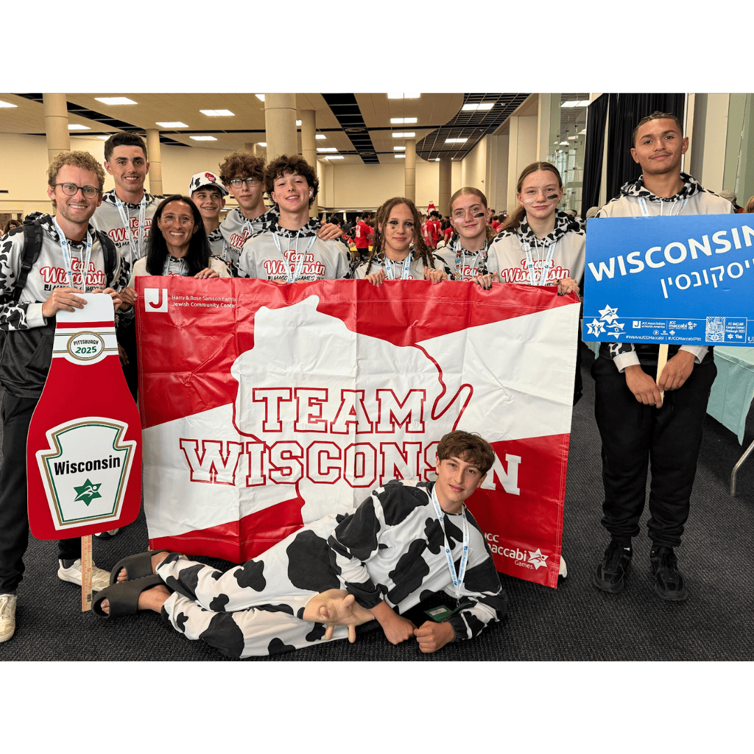 A group of young people pose indoors with a large "Team Wisconsin" flag, a Wisconsin sign, and one person dressed in a cow costume lying in front.