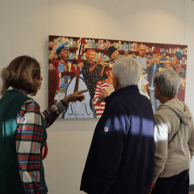 Three people stand closely together, looking at and discussing a colorful painting of musicians playing instruments displayed on a white wall.