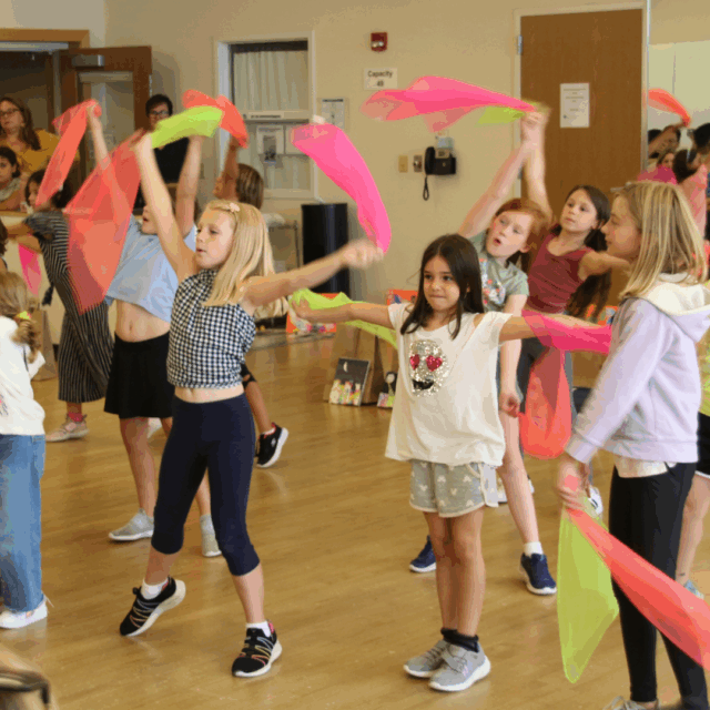 Children participate in a group dance activity indoors, holding colorful scarves and following along with arm movements.