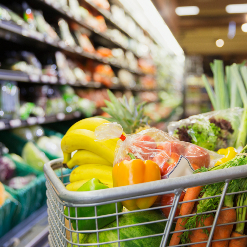 A shopping cart filled with various fresh produce like bananas, bell peppers, tomatoes, lettuce, and carrots in a grocery store produce aisle.