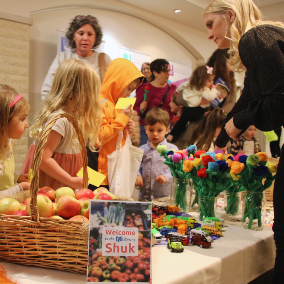 People, including children, gathered around a table with a basket of apples, colorful paper flowers, and toys. A sign on the table reads, "Welcome to the Library Shuk.