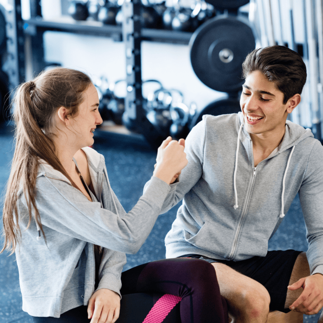 Two people sitting on the floor of a gym, smiling and fist bumping each other. They wear casual workout clothes, and gym equipment is visible in the background.