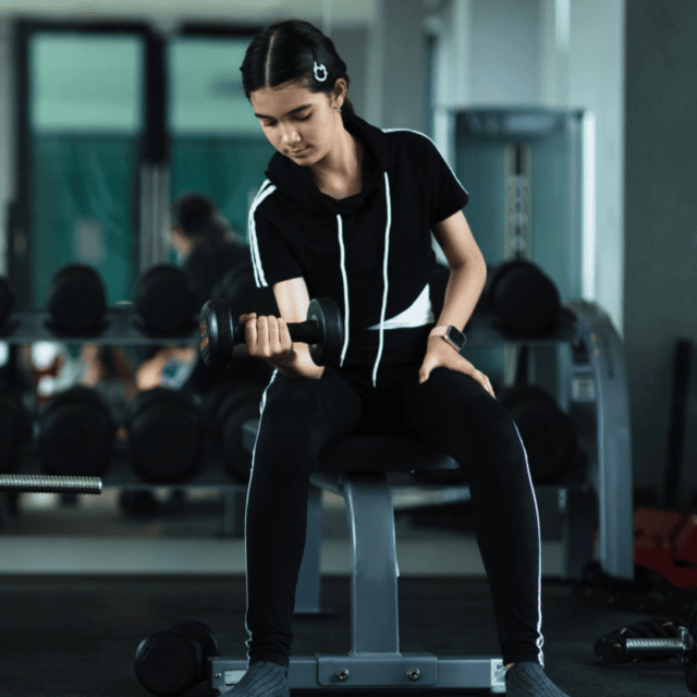 A person in a gym is sitting on a bench, lifting a dumbbell with one arm. They are wearing black athletic wear.