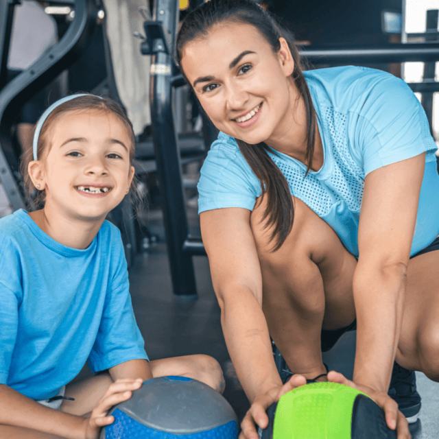 A woman and a girl are in a gym, both wearing blue shirts. They are kneeling on the floor and smiling, each holding a weighted exercise ball.