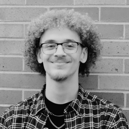A person with curly hair and glasses standing against a brick wall, wearing a checkered shirt and smiling slightly. (Black and white photo).