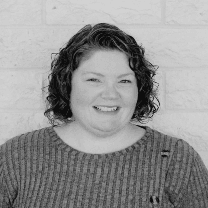 A person with curly hair smiles while wearing a striped sweater in front of a light-colored brick wall.