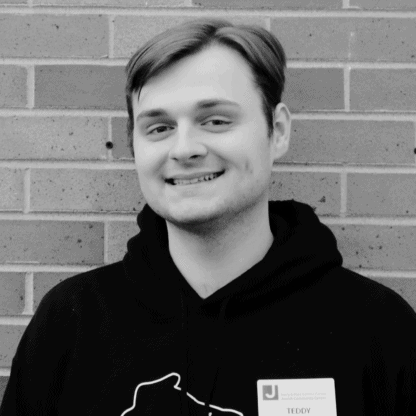 A person in a hoodie is smiling in front of a brick wall. They are wearing a name badge.