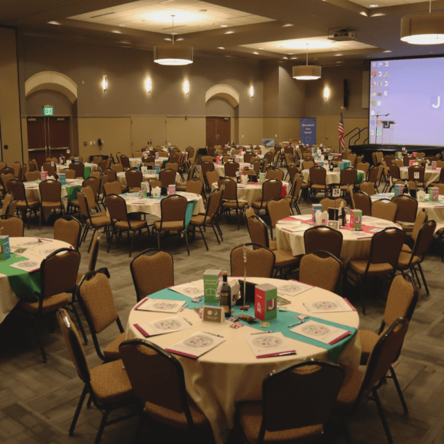 A conference room with round tables set for an event. Each table has papers, binders, and folders. A projector screen is visible in the background.