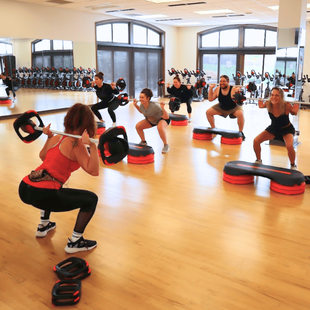 A group fitness class with six people holding weights and performing squats. They are in a gym with large mirrors and wooden floors.