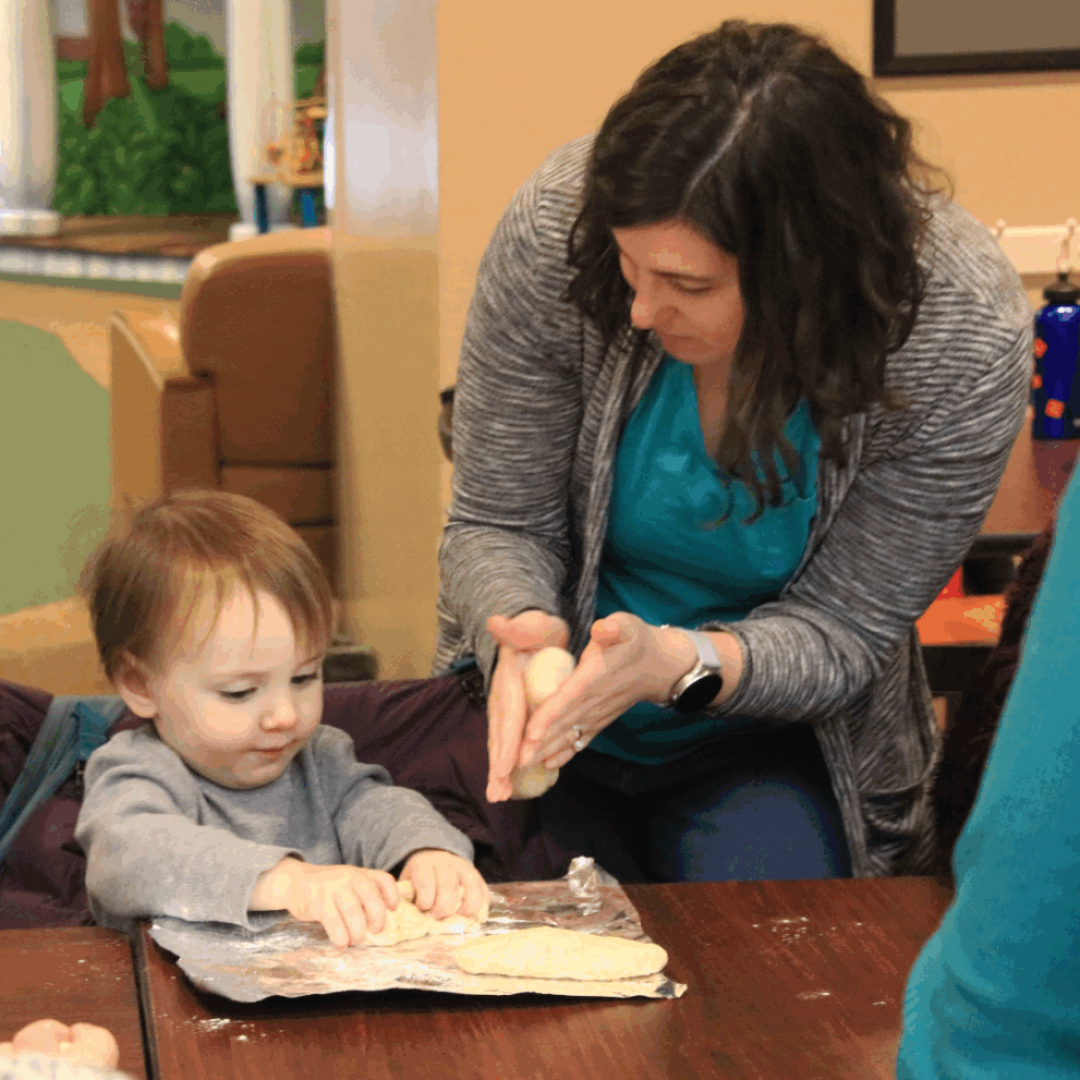 An adult helps a young child shape dough on a table covered with plastic wrap inside a room.