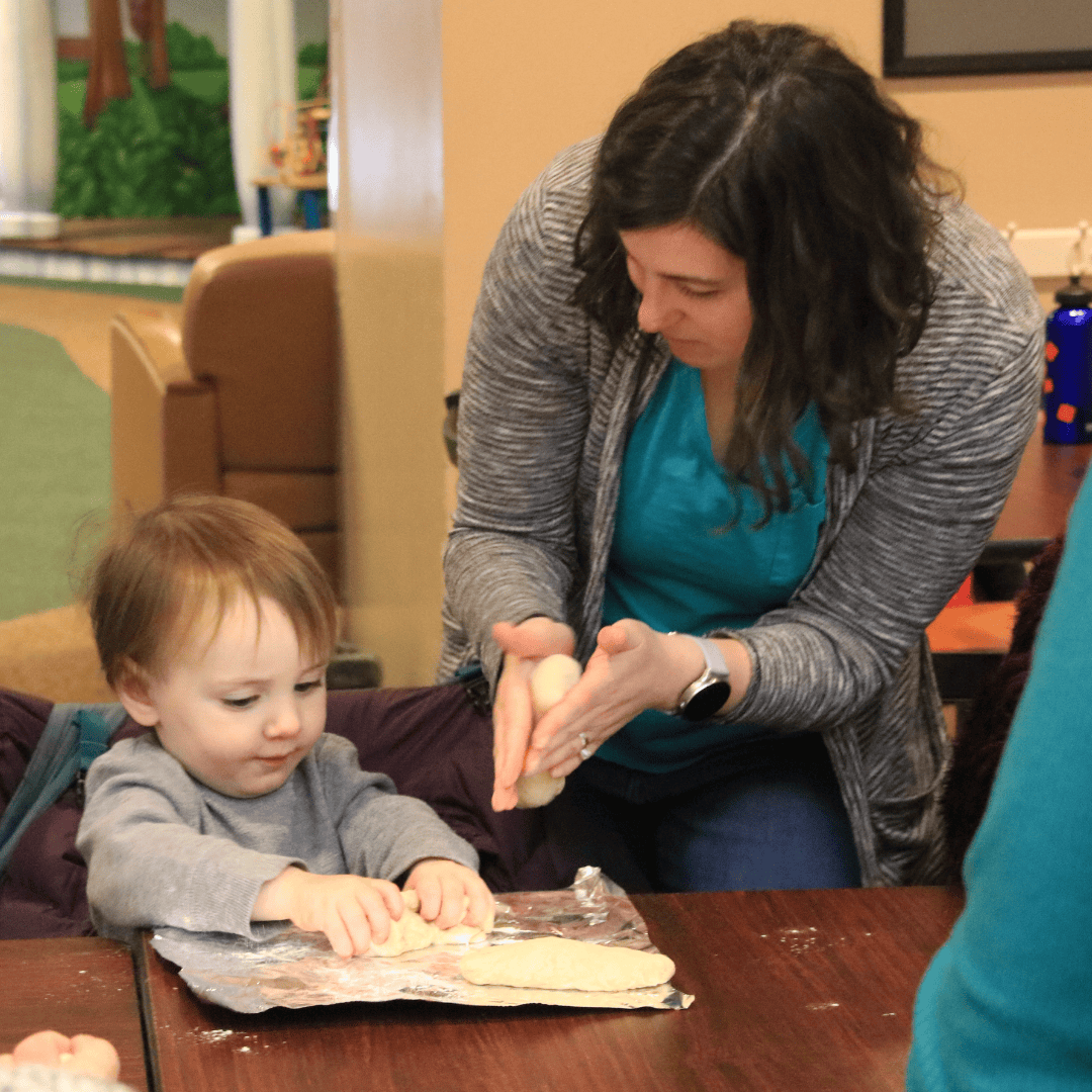 An adult helps a young child shape dough on a table covered with plastic wrap inside a room.