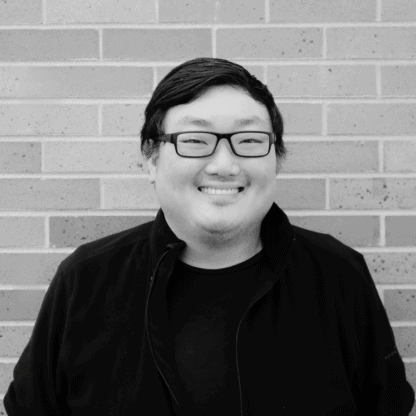 A person with short dark hair and glasses smiles while standing in front of a brick wall, wearing a black top.