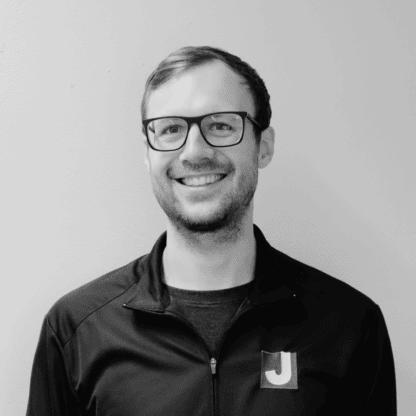 A man with glasses and a beard is smiling, wearing a zip-up jacket with a "J" logo, standing against a plain light-colored wall.