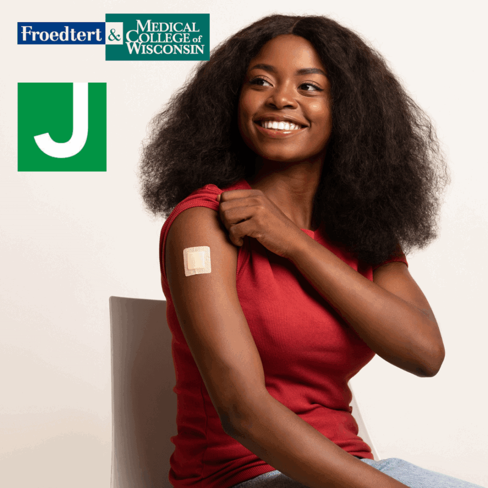 A smiling woman in a red shirt sits on a chair showing a bandage on her upper arm. Froedtert & Medical College of Wisconsin and a green "J" logo are visible in the corner.