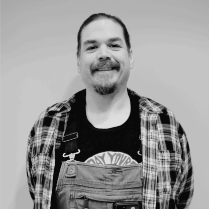 A man with a goatee and mustache smiles at the camera, wearing a plaid shirt over a graphic tee and overalls, standing against a plain background.