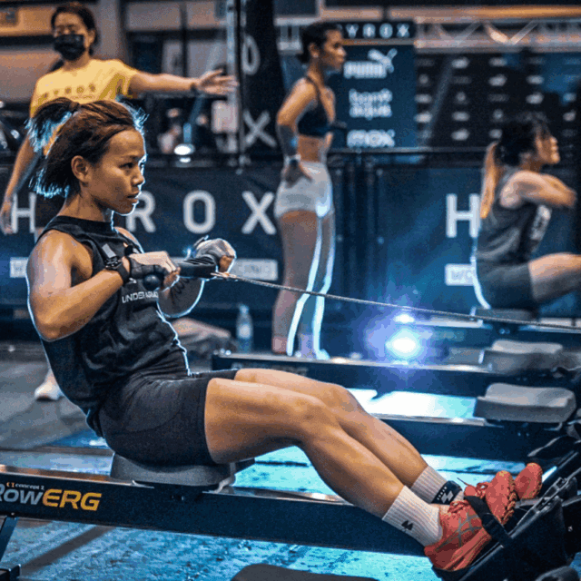 A woman rows on an indoor rowing machine during a fitness competition, with other participants and a judge visible in the background.