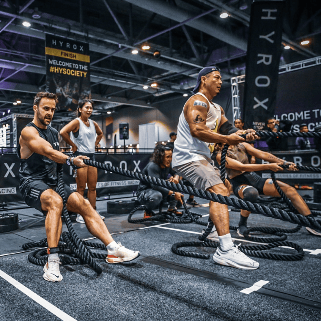 Several people participate in a competitive rope-pulling fitness event inside an indoor gym venue, with banners and spectators visible in the background.