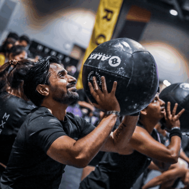 A man in athletic clothing lifts a large medicine ball during a fitness event, with other participants doing the same in the background.