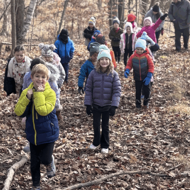 A group of children dressed in winter clothes walk along a leaf-covered trail in a forest, accompanied by an adult.