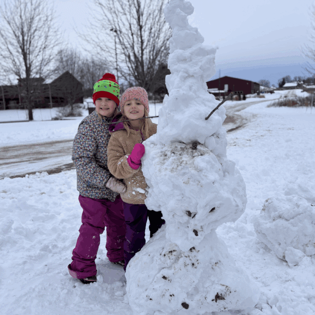 Two children in winter clothing stand beside a large, irregular snowman on a snowy ground with trees and buildings in the background.