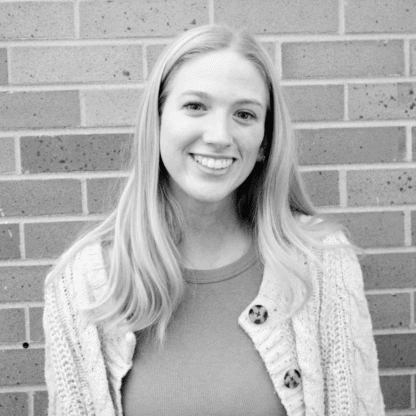 A young woman with long hair and a sweater smiles while standing in front of a brick wall.