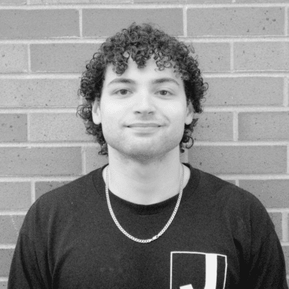 A young person with curly hair and a chain necklace stands in front of a brick wall, wearing a dark shirt with a white graphic on the chest.